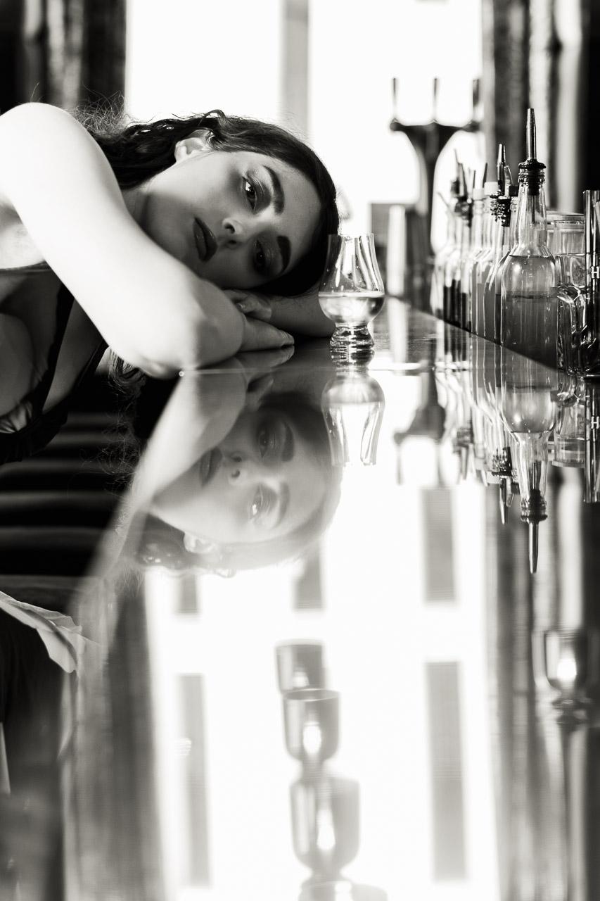 Absente Présence est un portrait en noir et blanc d’une femme perdue dans ses pensées, front posé sur un bar, face à un verre vide. Son reflet flou évoque l’attente, le manque, peut-être un rendez-vous manqué.