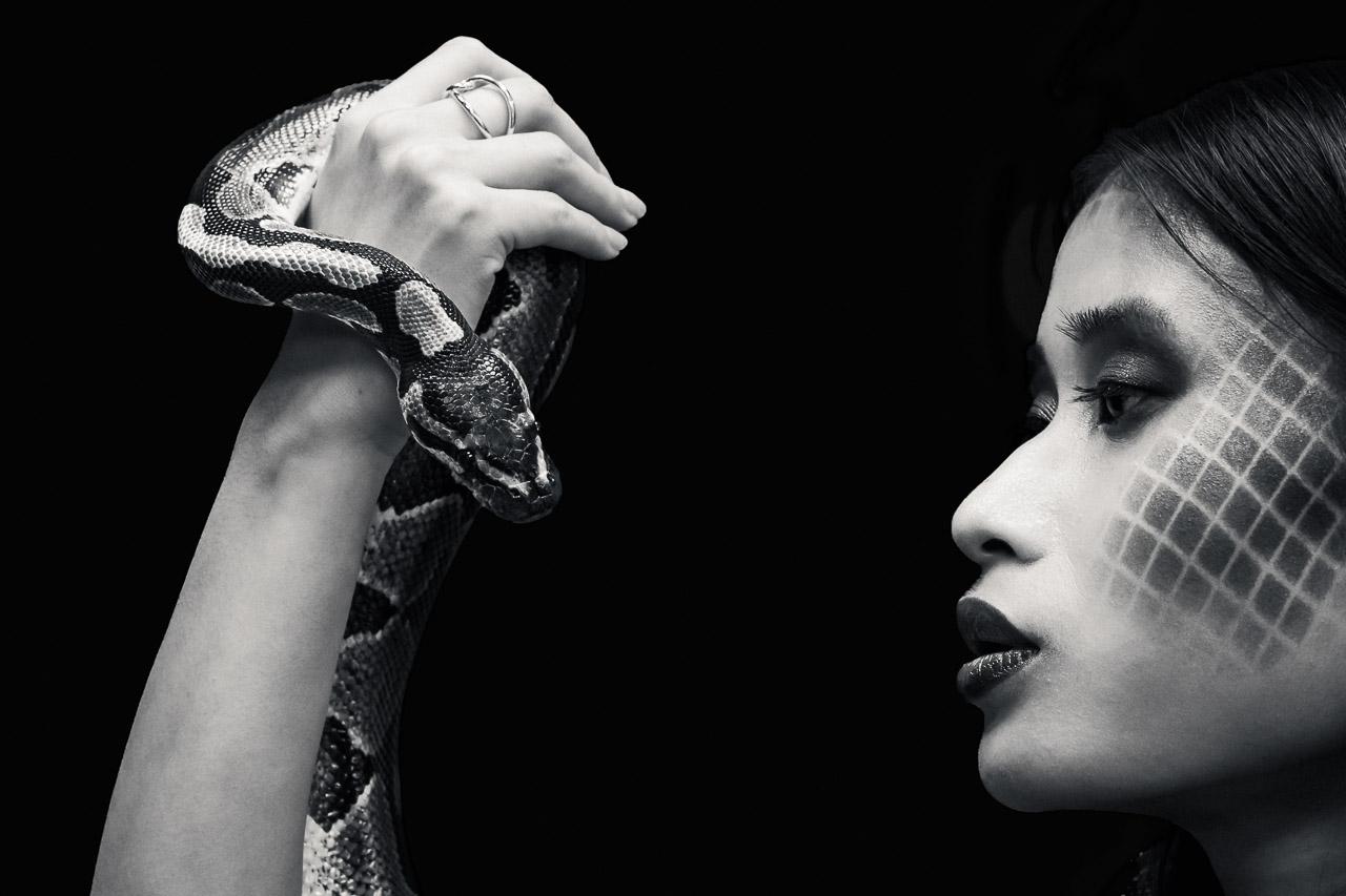 Reine Froide Portrait en noir et blanc d’une femme au maquillage géométrique faisant face à un serpent enroulé autour d’un bras, ambiance mystérieuse et symbolique.