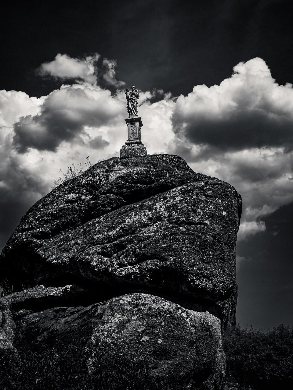 La Veille, photographie noir et blanc de Thierry Stentzel : statue de saint Joseph portant l’Enfant Jésus sur un immense rocher à Noirétable, sous un ciel nuageux.