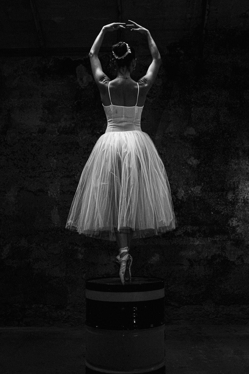 contrepoint Contrepoint : danseuse vue de dos, en tutu blanc et sur pointes, bras levés, sur un fût métallique devant un mur sombre, en noir et blanc.